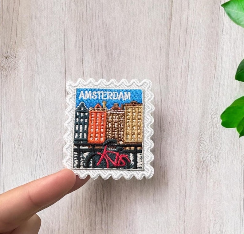 May include: A square embroidered patch with a scalloped white border featuring a blue sky, a row of colorful buildings in black, orange, brown, and tan, and a red bicycle in the foreground. The text AMSTERDAM is stitched in white at the top.