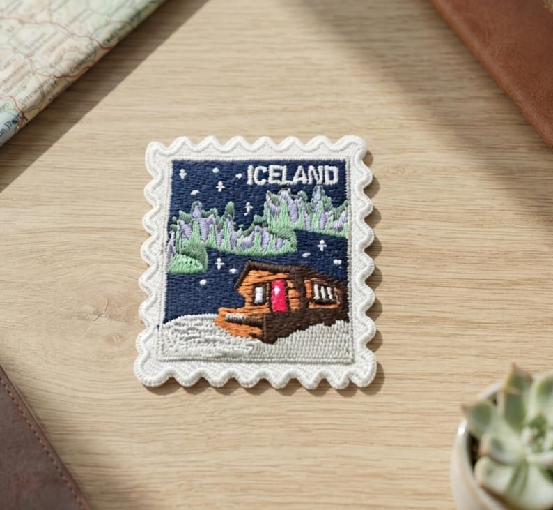 Pu&ograve; includere: Una toppa ricamata rettangolare a forma di francobollo con un bordo bianco smerlato. Il disegno raffigura una capanna marrone con una porta rosso brillante, un primo piano innevato e aurore boreali verdi su un cielo blu scuro con il testo ICELAND.