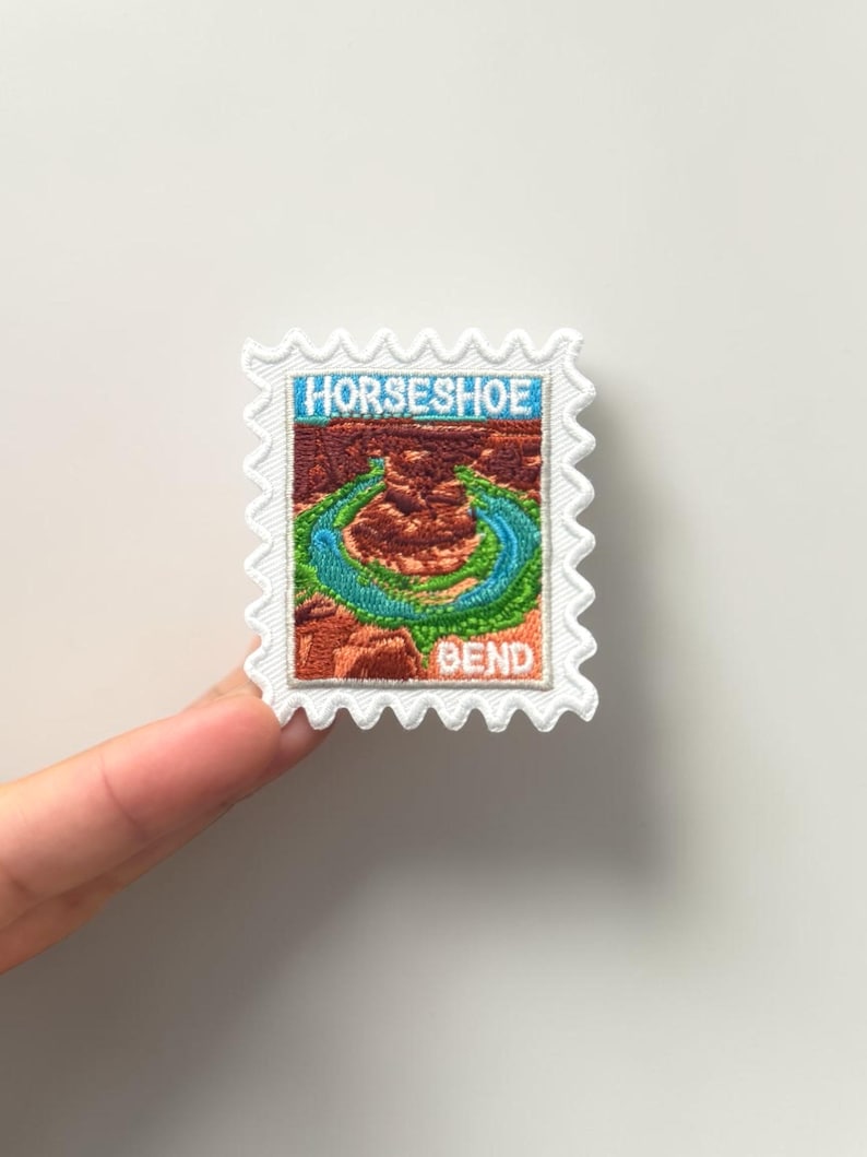 May include: A rectangular embroidered patch shaped like a postage stamp with a scalloped white border. The centre features a detailed landscape of Horseshoe Bend with a winding blue river, red rock formations, and the text HORSESHOE BEND.