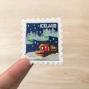 Pu&ograve; includere: Una toppa ricamata rettangolare con bordo bianco smerlato che raffigura un cielo notturno blu scuro, un'aurora boreale verde e una baita marrone con una porta rossa. La scritta ICELAND &egrave; ricamata in bianco nella parte superiore.