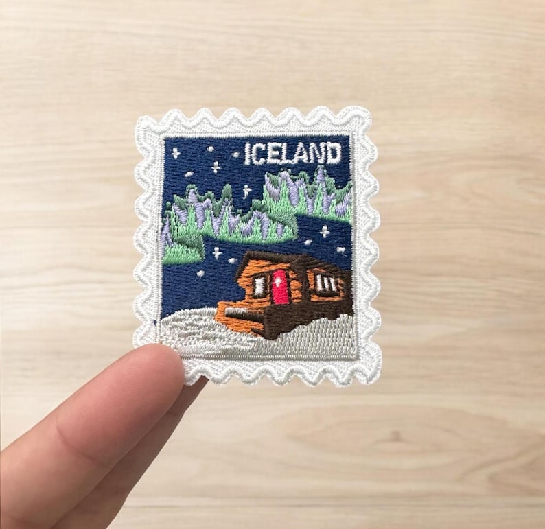 Pu&ograve; includere: Una toppa ricamata rettangolare con bordo bianco smerlato che raffigura un cielo notturno blu scuro, un'aurora boreale verde e una baita marrone con una porta rossa. La scritta ICELAND &egrave; ricamata in bianco nella parte superiore.