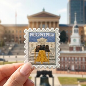 Pu&ograve; includere: Una toppa ricamata rettangolare con bordo bianco smerlato, raffigurante la Liberty Bell in giallo e marrone su una facciata di edificio beige e un cielo blu, con la parola PHILADELPHIA ricamata in bianco nella parte superiore.