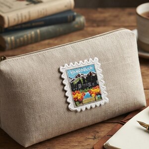 Puede incluir: Un estuche de lino beige con cremallera que presenta un parche bordado rectangular con forma de sello postal. El parche muestra un paisaje con monta&ntilde;as, &aacute;rboles y el texto Vancouver en hilo de colores azul, verde, naranja y blanco.