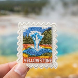 May include: A rectangular embroidered patch with a scalloped edge, featuring a colorful landscape scene of a geyser erupting. The design includes blue water, green trees, and orange ground, with the word YELLOWSTONE stitched in white at the bottom.