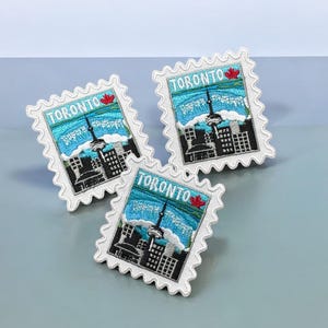 May include: Three rectangular embroidered patches shaped like postage stamps, each featuring a skyline of Toronto with the CN Tower, a blue sky, and a small red maple leaf. The text TORONTO is stitched in white across the top of each patch.