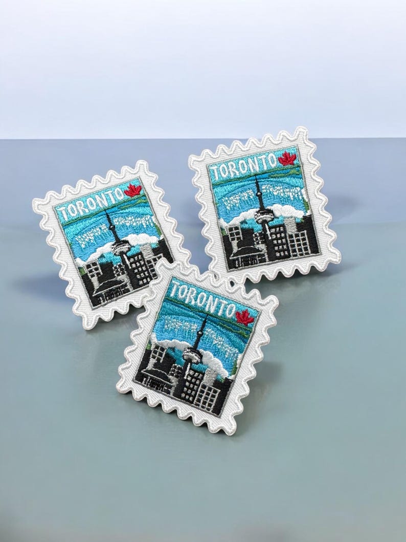May include: Three rectangular embroidered patches shaped like postage stamps, each featuring a skyline of Toronto with the CN Tower, a blue sky, and a small red maple leaf. The text TORONTO is stitched in white across the top of each patch.