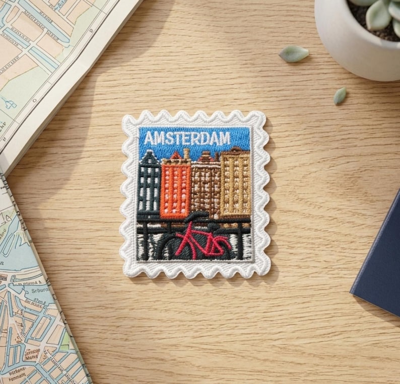 May include: A rectangular embroidered patch shaped like a postage stamp featuring a blue sky, a row of colorful buildings in shades of orange, brown, and tan, and a red bicycle in the foreground. The word AMSTERDAM is stitched in white letters.
