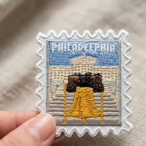 Pu&ograve; includere: Una toppa ricamata quadrata a forma di francobollo con un bordo bianco smerlato. Il design presenta un cielo azzurro sopra un edificio beige e una prominente campana gialla con dettagli neri, con la scritta PHILADELPHIA.