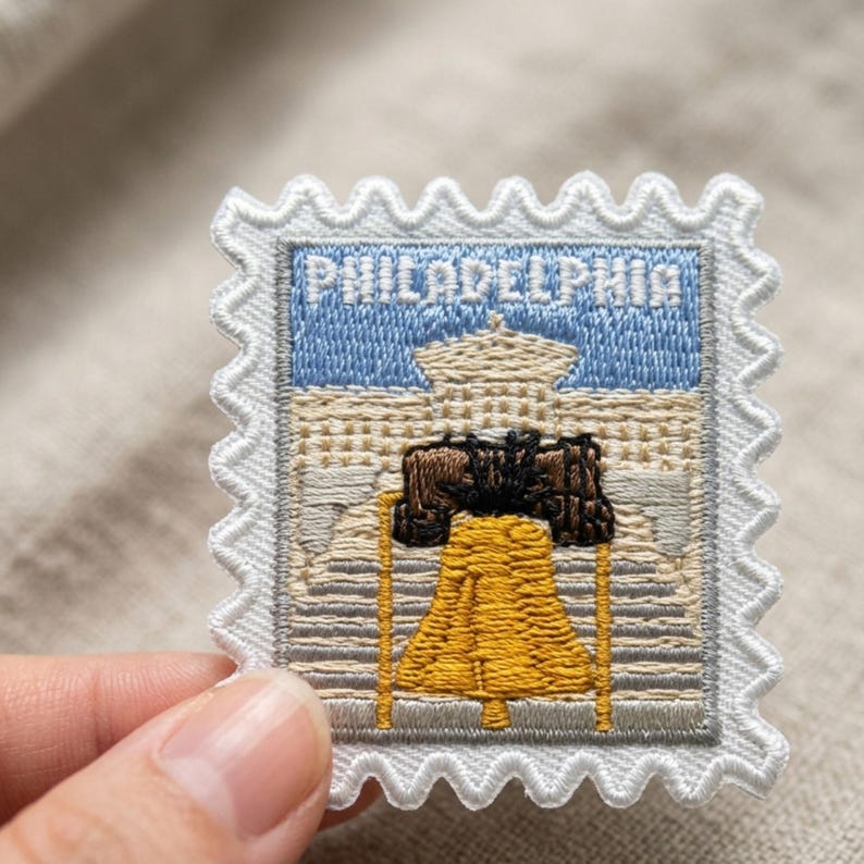 Pu&ograve; includere: Una toppa ricamata quadrata a forma di francobollo con un bordo bianco smerlato. Il design presenta un cielo azzurro sopra un edificio beige e una prominente campana gialla con dettagli neri, con la scritta PHILADELPHIA.