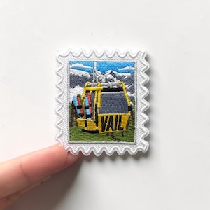 May include: Embroidered patch in a stamp design featuring a yellow ski gondola with the word "VAIL" on it. The patch has a white border with a scalloped edge. The background shows a mountain scene with blue sky.