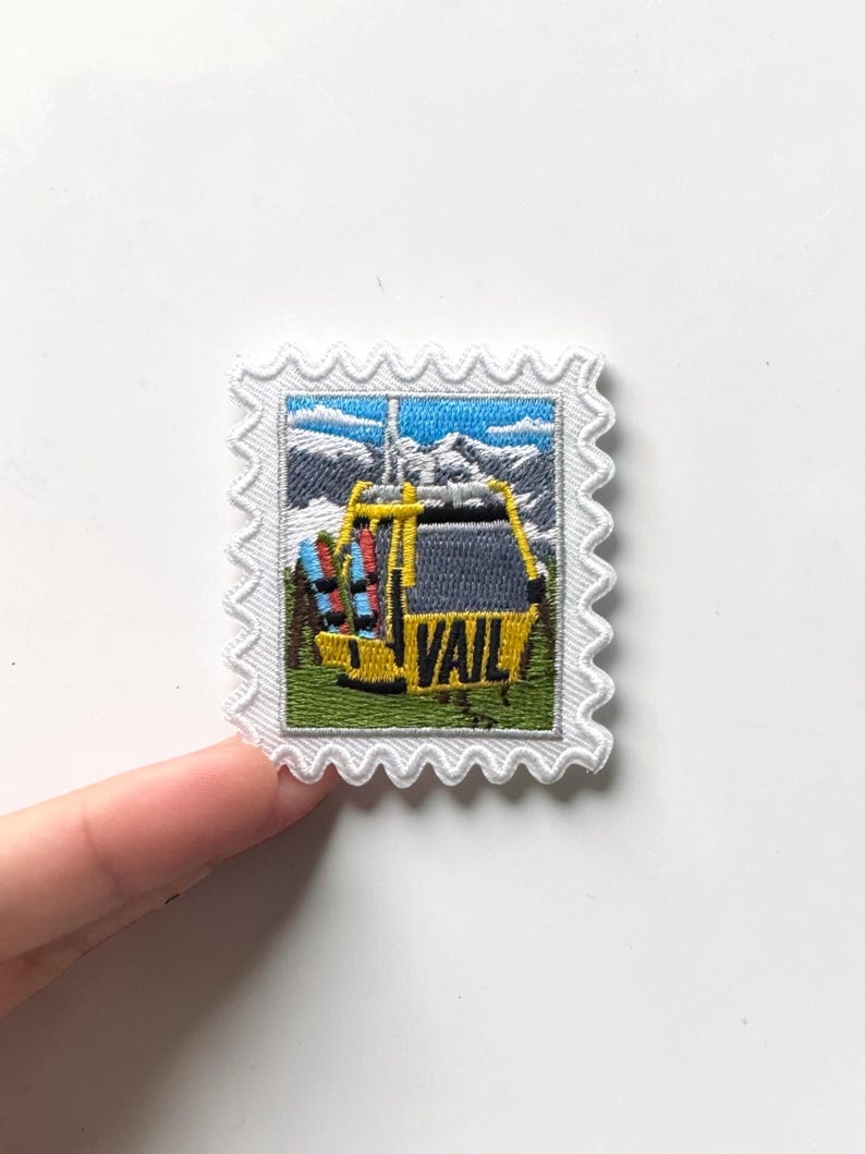May include: Embroidered patch in a stamp design featuring a yellow ski gondola with the word "VAIL" on it. The patch has a white border with a scalloped edge. The background shows a mountain scene with blue sky.