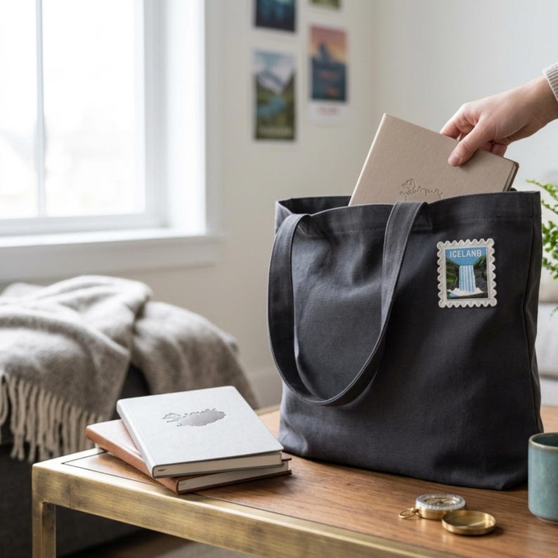 May include: A dark gray canvas tote bag featuring a rectangular patch with a scalloped edge depicting a waterfall and the text ICELAND. The bag has two shoulder straps and is shown with two stacked notebooks and a compass on a wooden table.