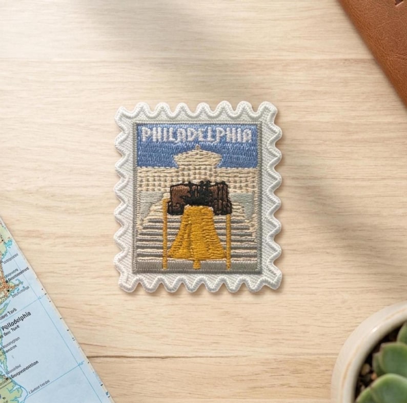 Pu&ograve; includere: Una toppa ricamata rettangolare a forma di francobollo raffigurante la Liberty Bell in giallo e marrone, su sfondo di un edificio beige e cielo blu, con la scritta PHILADELPHIA ricamata in bianco nella parte superiore del disegno.