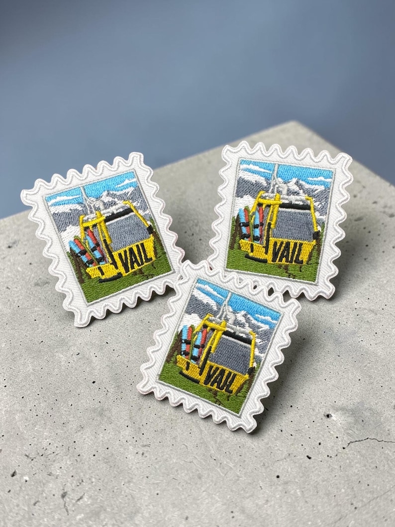 May include: Three embroidered patches in the shape of postage stamps. Each patch features a yellow ski lift with the word "VAIL" and a mountain scene. The patches have a white border with a scalloped edge.