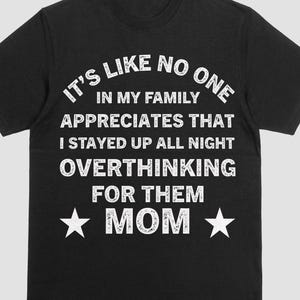 Peut inclure: T-shirt noir avec texte blanc : "IT'S LIKE NO ONE IN MY FAMILY APPRECIATES THAT I STAYED UP ALL NIGHT OVERTHINKING FOR THEM MOM" avec des étoiles. Le t-shirt a un col rond et des manches courtes.