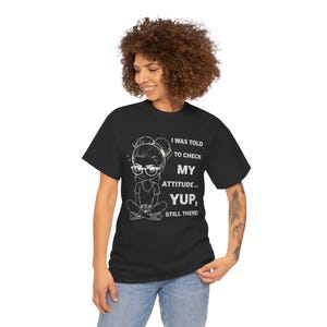 Peut inclure: T-shirt noir avec un dessin blanc d'une fille de dessin animé portant des lunettes et le texte "I WAS TOLD TO CHECK MY ATTITUDE... YUP, STILL THERE!". Le t-shirt est porté avec un jean bleu.