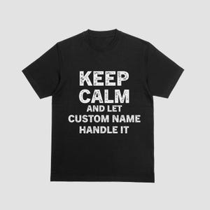 Könnte beinhalten: Schwarzes T-Shirt mit dem weißen Text "KEEP CALM AND LET CUSTOM NAME HANDLE IT". Der Text ist in einer kräftigen, verwitterten Schriftart gehalten. Das Shirt hat kurze Ärmel und einen Rundhalsausschnitt.