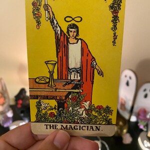 May include: A tarot card depicting The Magician. The card has a yellow background with a figure in red robes holding a wand. A table with a chalice and other items is in front of the figure. The card's title, "THE MAGICIAN," is at the bottom.