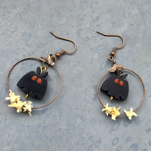 May include: A pair of dangle earrings featuring copper-toned circular hoops. Each hoop suspends a black moth-shaped charm with two orange eyes and a cluster of small, off-white, textured bone-like segments attached to the bottom of the wire hoop.