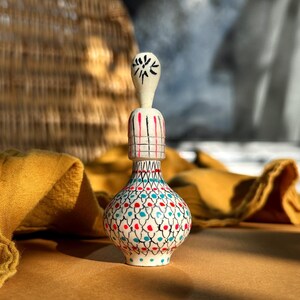May include: A white ceramic bottle with a blue, red and black geometric pattern. The bottle has a small, round, white stopper with a black star design.
