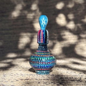 Pode incluir: Uma garrafa de cerâmica azul e branca com um design decorativo. A garrafa tem uma pequena rolha pontiaguda e é decorada com padrões geométricos coloridos.