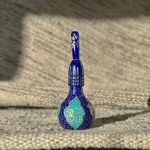 Pode incluir: Pequena garrafa azul ornamentada, com um pescoço longo e fino e um fecho decorativo. A garrafa apresenta um design floral turquesa e dourado no corpo, com cerca de 10 cm de altura.