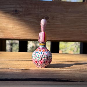 Pode incluir: Uma garrafa decorativa pintada à mão, com uma base rosa e um padrão floral detalhado em azul, vermelho e amarelo. A garrafa tem um pescoço longo e fino com uma rolha rosa. O design é intrincado e colorido, mostrando um estilo de arte popular.