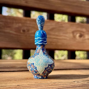 Pode incluir: Pequena garrafa azul decorativa com tampa a condizer. A garrafa apresenta um padrão floral detalhado em tons de azul, vermelho e branco. A tampa é azul clara com salpicos brancos. A garrafa tem cerca de 10 cm de altura.
