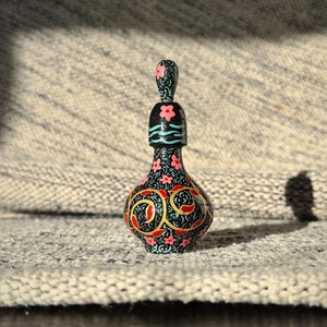 Puede incluir: Una botella negra pintada a mano con un diseño floral. La botella tiene un cuerpo pequeño y redondo y un cuello largo y delgado. Las flores son rosas y rojas, y las hojas son verdes. La botella está decorada con detalles dorados.
