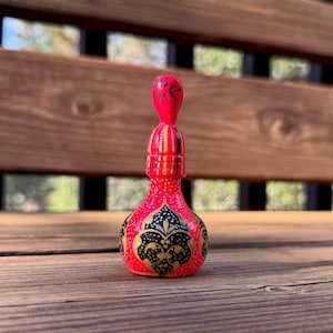 Pode incluir: Uma pequena garrafa decorativa vermelha com um pescoço longo e fino e uma base arredondada. A garrafa apresenta intrincados padrões florais pretos e dourados e uma rolha vermelha a condizer. A garrafa é exibida numa superfície de madeira.