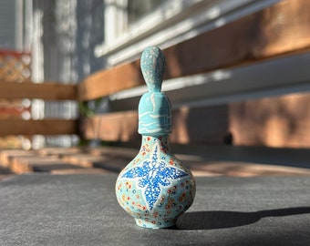Puede incluir: Una pequeña botella de cerámica azul con un diseño floral blanco y rojo. La botella tiene un tapón con un diseño blanco y azul.