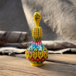 May include: A hand-painted, decorative wooden bottle with a yellow base and top. The bottle's body is adorned with colorful floral patterns in red, blue, and pink. The bottle is approximately 5 inches tall and sits on a wooden surface.