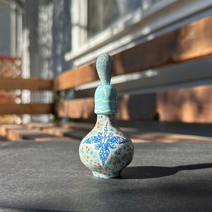 May include: A small, blue ceramic bottle with a white and red floral design. The bottle has a stopper with a white and blue design.