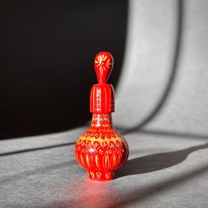 Könnte beinhalten: Eine rote, verzierte Flasche mit dekorativem Verschluss. Die Flasche weist aufwendige gelbe und grüne Blumenmuster auf. Das Design ist symmetrisch, mit einem bauchigen Boden und einem schlanken Hals. Der Verschluss hat ein stilisiertes Blumenmuster.