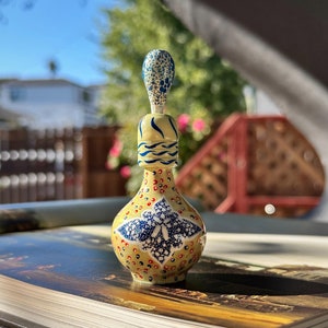 Pode incluir: Uma pequena garrafa de cerâmica amarela com um design azul e branco. A garrafa tem um corpo redondo e um pescoço longo e fino. A garrafa é decorada com pequenos pontos brancos e um padrão floral azul e branco.