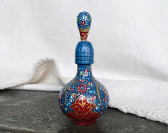Puede incluir: Una pequeña botella de vidrio azul pintada a mano con un diseño floral en rojo, dorado y blanco. La botella tiene un tapón con una parte superior bulbosa.