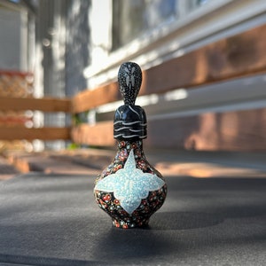 Pode incluir: Uma pequena garrafa de cerâmica pintada à mão com um design floral preto e branco. A garrafa tem um corpo redondo e um pescoço alto e fino com uma rolha preta e branca. A garrafa é decorada com uma variedade de cores, incluindo vermelho, azul e dourado.