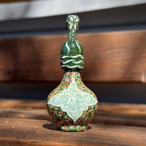 May include: A decorative, hand-painted bottle with a bulbous base and a long, slender neck. The bottle features a green, gold, red, and white floral design. The bottle is approximately 15 cm tall.