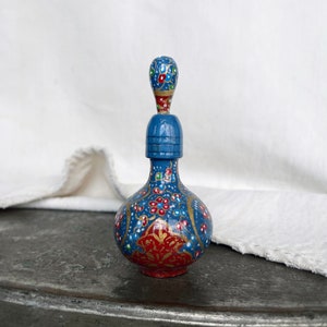 Könnte beinhalten: Eine kleine, handbemalte blaue Glasflasche mit einem floralen Design in Rot, Gold und Weiß. Die Flasche hat einen Stopfen mit einem bauchigen Oberteil.