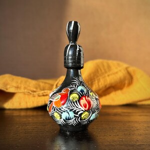 Pode incluir: Uma pequena garrafa preta com uma rolha branca e preta. A garrafa é decorada com desenhos florais e de pássaros coloridos.