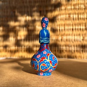 Pode incluir: Uma pequena garrafa de vidro azul pintada à mão com um desenho floral vermelho e dourado. A garrafa tem um tampão com flores cor-de-rosa.