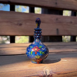 Pode incluir: Pequena garrafa de vidro azul ornamentada com um desenho floral. A garrafa apresenta uma flor vermelha com folhas verdes e uma rolha escura e bulbosa. A garrafa está sobre uma superfície de madeira.