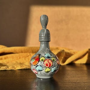 Pode incluir: Uma garrafa de cerâmica cinza pintada à mão com uma rolha decorativa. A garrafa apresenta desenhos florais e de pássaros coloridos em vermelho, laranja, amarelo e azul. A garrafa é exibida em uma superfície de madeira escura.