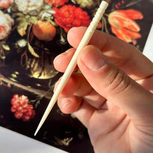Puede incluir: Primer plano de un palillo de dientes de madera blanco con un dise&ntilde;o texturizado y estriado. El palillo de dientes se sostiene en una mano sobre un fondo de una pintura de flores.