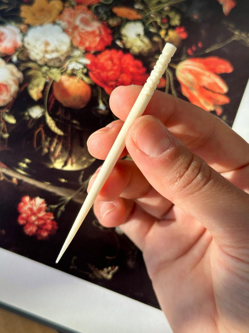 Puede incluir: Primer plano de un palillo de dientes de madera blanco con un dise&ntilde;o texturizado y estriado. El palillo de dientes se sostiene en una mano sobre un fondo de una pintura de flores.