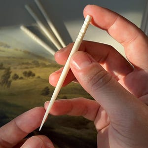 Puede incluir: Primer plano de una mano sosteniendo un palillo de dientes de madera blanco. El palillo de dientes tiene una superficie texturizada y un extremo puntiagudo.
