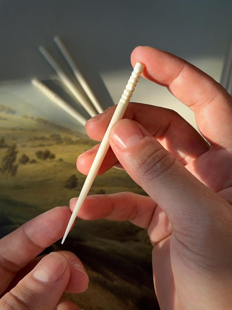 Puede incluir: Primer plano de una mano sosteniendo un palillo de dientes de madera blanco. El palillo de dientes tiene una superficie texturizada y un extremo puntiagudo.