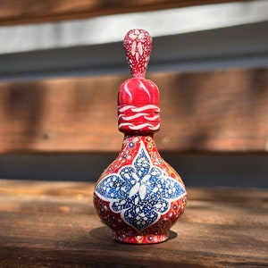 Pode incluir: Uma pequena garrafa de cerâmica pintada à mão com um desenho floral vermelho, branco e azul. A garrafa tem um corpo redondo e um pescoço estreito com uma rolha.