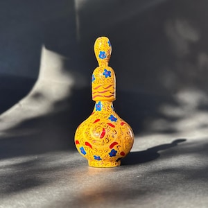 Puede incluir: Una botella de vidrio amarilla con un diseño floral en rojo, azul y dorado. La botella tiene un cuello estrecho y un cuerpo redondo.