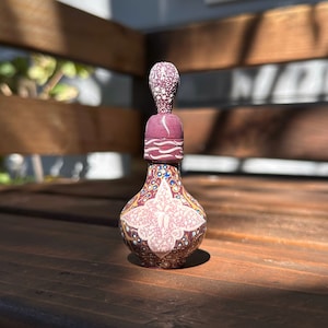 Pode incluir: Uma pequena garrafa de cerâmica pintada à mão com um rolha decorativo. A garrafa é decorada com um desenho floral rosa e branco, e a rolha é decorada com pontos brancos. A garrafa está sentada sobre uma superfície de madeira.